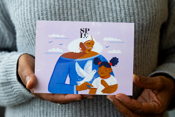 An image of a Black woman’s hands holding a lavender card with artwork depicting a Black elder and child embracing a dove of peace.